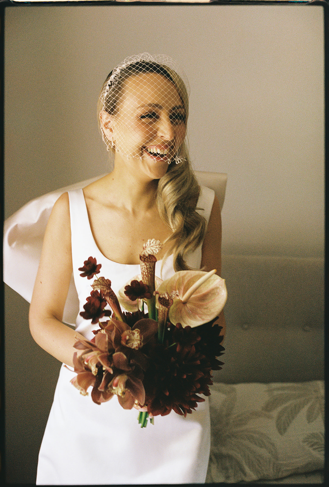 miling bride wearing a custom silk wedding gown with back bow and birdcage veil, holding a burgundy and blush bridal bouquet by The Beautiful Bunch – Melbourne winery wedding at Marnong Estate photographed by Kinship by Kristy