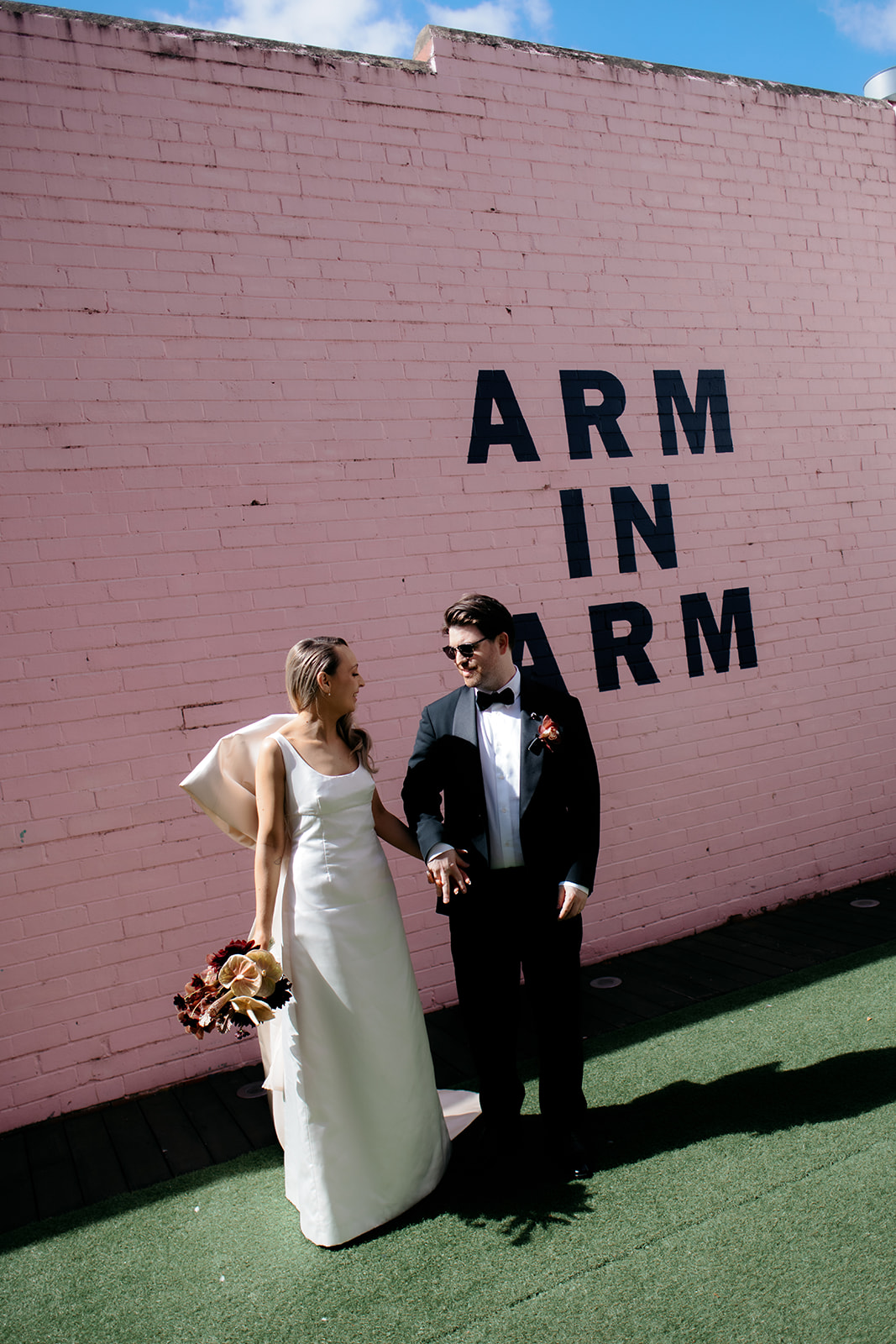 Bride and groom holding hands in front of a pink "Arm in Arm" wall mural in Northcote, Melbourne – bride wearing custom silk wedding gown with back bow, groom in black tuxedo – wedding photography by Kinship by Kristy