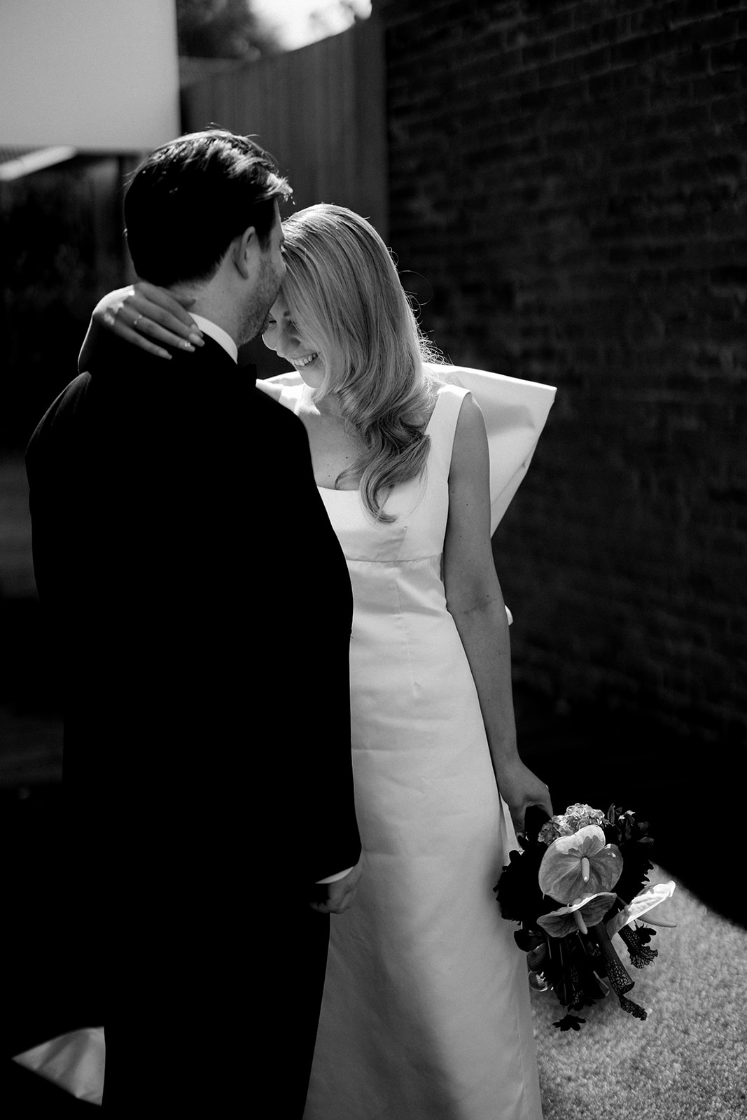 Black and white romantic couple portrait of bride and groom laughing together in a laneway in Northcote, Melbourne — bride in custom-designed silk gown holding burgundy bridal bouquet by The Beautiful Bunch, photography by Kinship by Kristy